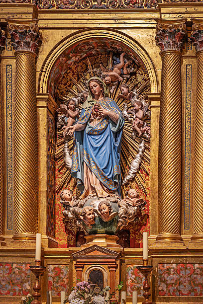 Iglesia de Santo Tome, Toledo, Spain, Europe, September 16, 2025: Editorial image of a side altar inside Iglesia de Santo Tomé in Toledo, Spain, featuring a statue of the Virgin Mary dressed in blue and white robes with gold accents. The figure stands on a cloud supported by cherubic heads, framed by radiant golden rays and surrounded by ornate gilded columns and decorative elements. The altar includes candles and floral arrangements at the base, enhancing the ceremonial and sacred atmosphere. The composition reflects traditional Catholic iconography and Spanish baroque craftsmanship
