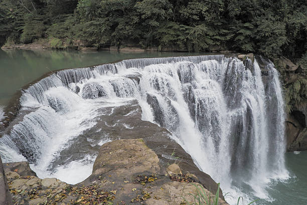 istock A scenic view of a wide waterfall cascading over multiple rock formations into a calm river below, surrounded by lush green forest. 2239881114