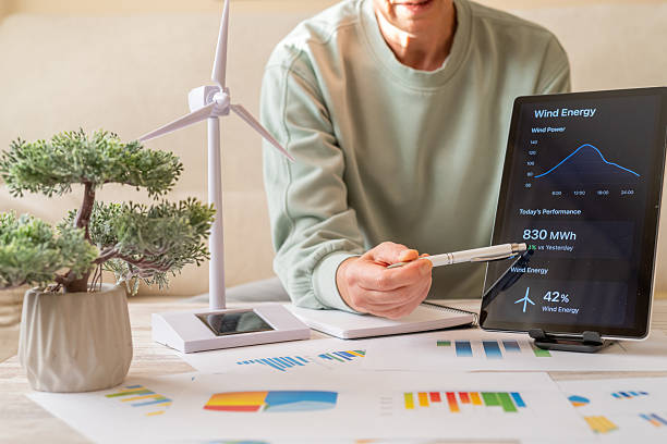 istock Person calculating sustainable wind power statistics using a digital tablet application, with a miniature wind turbine and chart documents on the desk, representing green energy management 2239883998