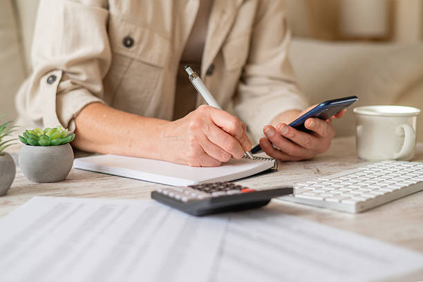 istock Woman's hands writing notes in a spiral notebook while holding a smartphone, actively managing personal finances, calculating a budget, and planning household expenses on a desk 2239884058