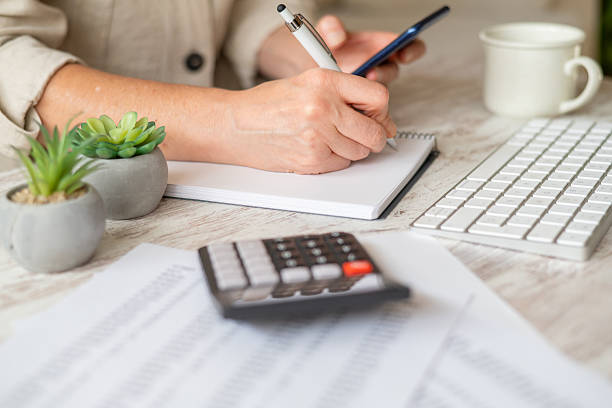 istock Person is calculating finances and managing household expenses, writing in a notebook while using a smartphone, with a calculator, documents, and keyboard on a desk 2239884077