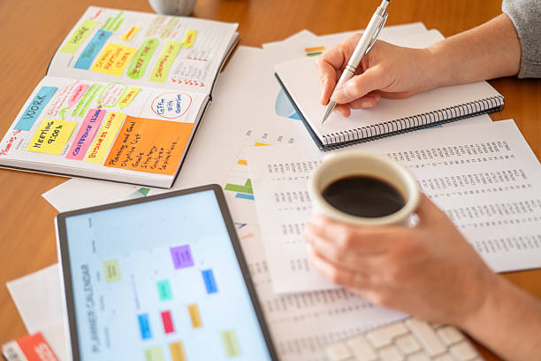 istock Person organizing work and home tasks in a planner, writing notes, and reviewing financial data on paper documents and a digital tablet, while drinking a hot cup of coffee 2239884291
