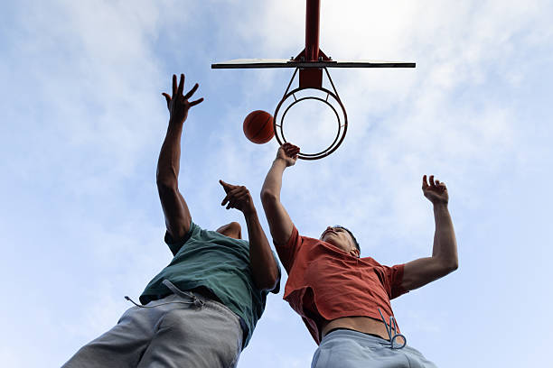 A low-angle wide view of two teenage boys playing basketball in a public basketball court in Newcastle, England. They are wearing sports clothing on an overcast day. They are playing a 1v1 basketball game, taking turns attacking and defending.Videos are available similar to this scenario.