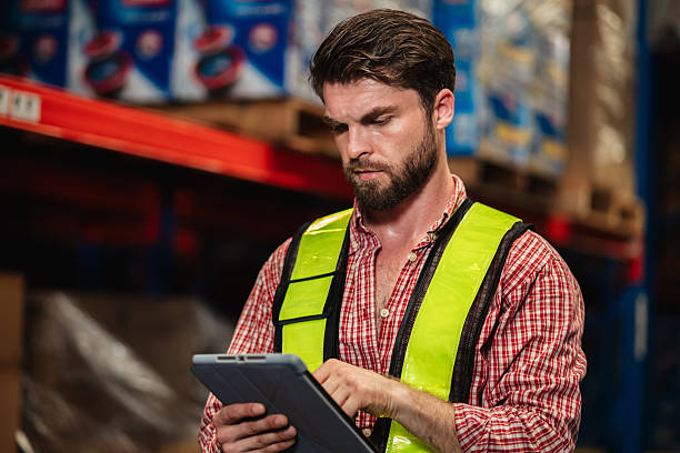 Warehouse workers and manager in reflective vests inspecting inventory, using digital tablet for stock control, logistics, supply chain management, and modern industrial warehouse operations.