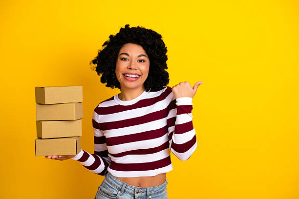 istock Young woman holding cardboard boxes and smiling confidently while pointing at them against a bright yellow background 2239896998