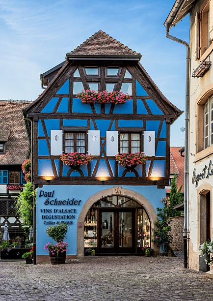 Eguisheim, France – June 16, 2025: Street view in Eguisheim, Alsace, France, featuring a pink half-timbered house with blue shutters, flowers, and climbing plants.