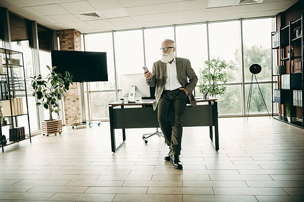 istock Mature businessman standing in stylish office environment confidently using his smartphone, representing leadership and elegance. 2239898626