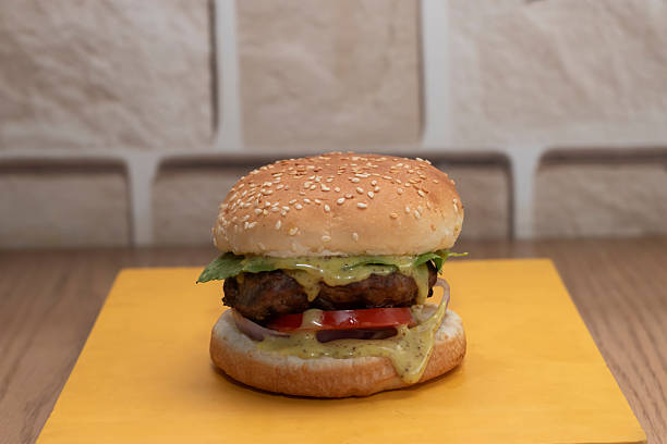 istock Basic Beef Burger with Sesame Seed Bun, Fresh Vegetables, patty, fresh lettuce, red onion, tomato and Creamy Herb Sauce on a yellow mat with a blurred brick wall background 2239902133