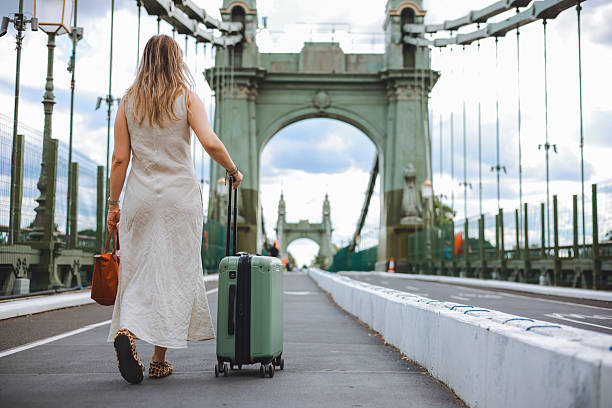 Woman walks confidently across a city bridge, pulling a green suitcase behind her. She is dressed in a floral top and white dress, enjoying her urban adventure with historic architecture and city life in the background, creating a vibrant travel scene.