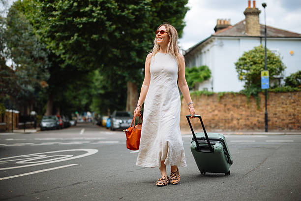 A happy adult black female entrepreneur on business travel in London. She is holding the handle of her large suitcase while standing on a sidewalk waiting for her taxi to take her to her hotel. She has an afro hairstyle and is wearing all black.