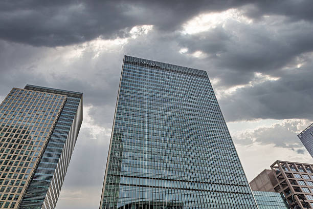 Upward view of a modern JPMorgan glass skyscraper beneath dramatic storm clouds and sun rays. London, UK, 14 May 2023