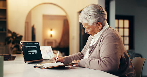istock Senior woman, writing and elearning with laptop for online education, research or course in home. Elderly, female person or taking notes with book or computer at night for web or distance learning 2239920024