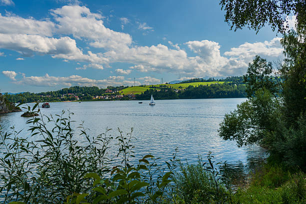 White sailboat glides peacefully across calm lake water surrounded by green hills and summer clouds. Perfect view of freedom, adventure, and nature recreation for travel and outdoor lifestyle concepts.
