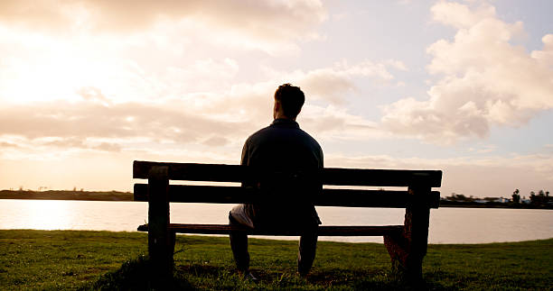 istock Man, thinking or back view with sunset on bench at lake for daydreaming, memory or break in nature. Male person, relax and fresh air with cloudy sky for rest, contemplating or travel at river or pond 2239924522