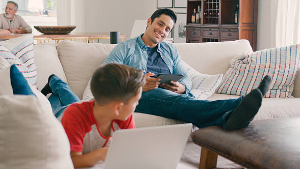 istock Father Sitting On Sofa At Home With Digital Tablet As Son Uses Laptop 2239925342