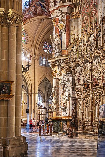 Catedral Primada de Santa Maria, Toledo, Spain, Europe, September 16, 2025: Interior view of the Catedral Primada de Santa María in Toledo, Spain, highlighting a richly sculpted wall adorned with religious figures and biblical reliefs. The Gothic architecture features vaulted ceilings, stone columns, and stained glass windows casting colored light into the space. A few visitors stand near the central column, providing scale and context to the cathedral’s vast interior.