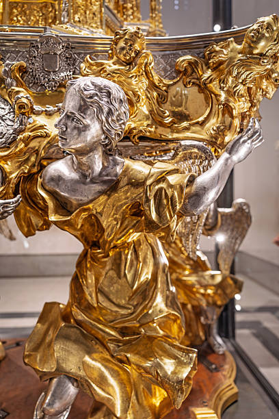 Catedral Primada de Santa Maria, Toledo, Spain, Europe, September 16, 2025: Close-up view of a richly adorned angelic figure inside the Catedral Primada de Santa María in Toledo, Spain. The sculpture, crafted from silver and gold, depicts a serene angel with flowing hair and textured wings, dressed in a gilded robe with elaborate folds. Surrounding the figure are ornate Baroque decorations, including cherubic faces and swirling scrollwork, emphasizing divine presence and ceremonial grandeur.