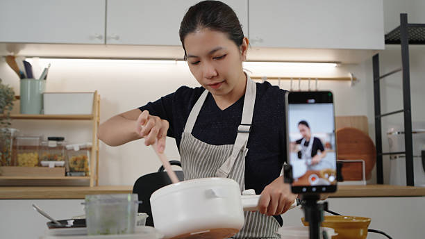 istock Asian woman in apron stirs ingredients while streaming a cooking vlog at home kitchen, smiling with confidence as an online influencer sharing recipes. 2239930337