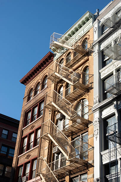 Sunlit view of historic New York City apartment buildings featuring ornate architectural details and metal fire escapes. The warm tones of the brickwork and contrasting window trims are illuminated against a clear blue sky, capturing the charm and texture of classic urban architecture.
