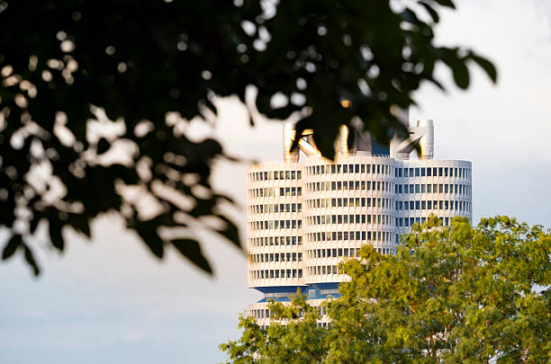 Contemporary high-rise building with distinctive cylindrical design, seen through tree leaves and park greenery. Warm natural light highlights the architectural details in a balanced urban scene.