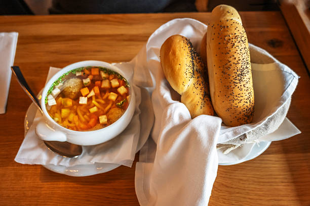 Soup in bowl (strong beef broth with vegetable cut into cubes with dumpling) and two bun with poppy seed in basket with white towel on wooden table.