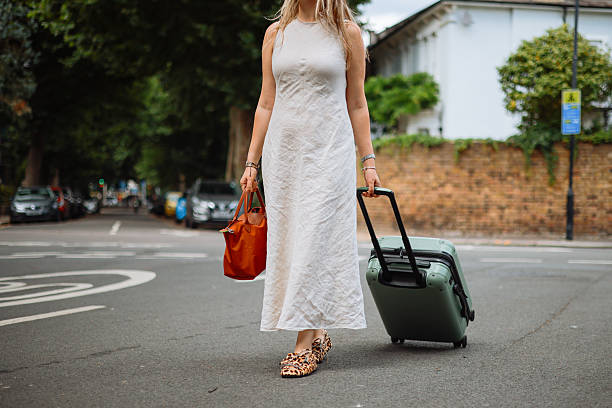 A happy adult black female entrepreneur on business travel in London. She is holding the handle of her large suitcase while standing on a sidewalk waiting for her taxi to take her to her hotel. She has an afro hairstyle and is wearing all black.