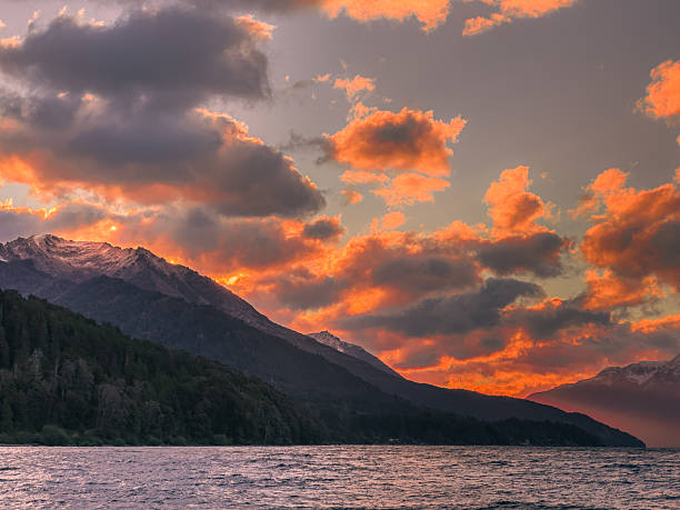Dramatic sunset over Lake Traful, with fiery clouds, sun rays illuminating the water, and snow-capped mountains in the background