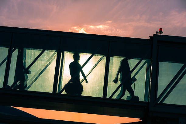 Group of passengers with luggage walking through boarding bridge to airplane at airport. Silhouettes of unrecognizable people at sunset.