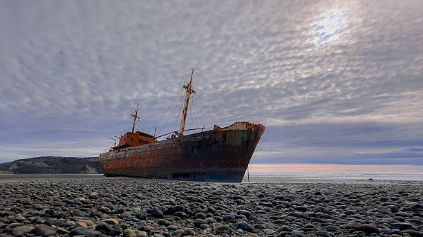 Rusty decaying shipwreck of the Desdemona, beached on a pebble and sand shore in Tierra del Fuego. The large vessel, with its mast and superstructure still intact, dominates the frame under a dramatic, cloudy sky with a hint of sun breaking through. The ocean stretches out to the right, and distant cliffs are visible on the left, creating a poignant scene of maritime history and the raw beauty of the Patagonian coast