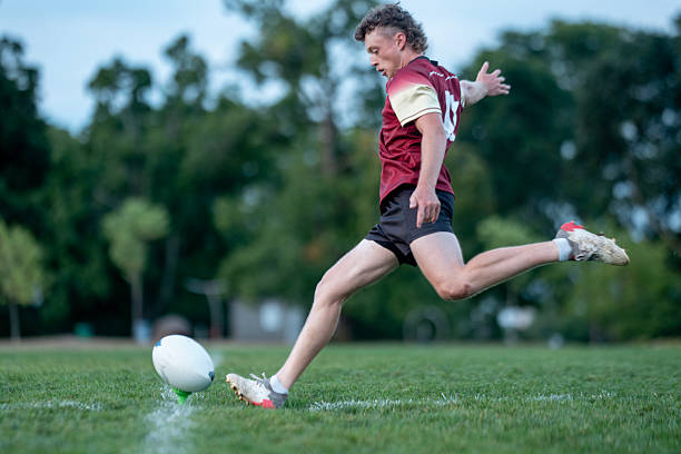 A Caucasian male rugby player in maroon and cream kit charges forward to kick a rugby ball on a sunny green field, capturing athletic momentum and the high-energy spirit of team sport.