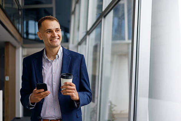 istock Smiling businessman walking in modern office, holding coffee cup and smartphone, having a break 2239969987