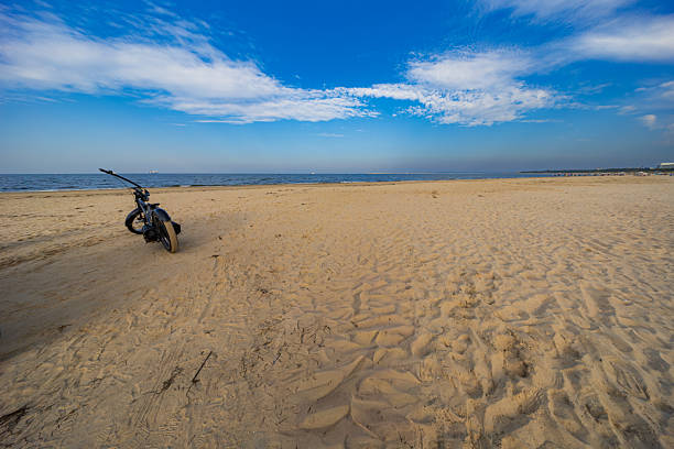 Serene Baltic Sea beach featuring an intriguing, unusually shaped black bicycle on vast golden sands under a clear blue sky with sparse clouds
