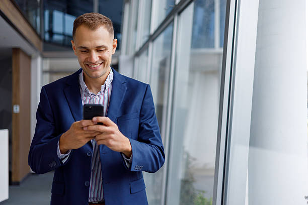 istock Businessman smiling, texting on phone, standing in modern office building corridor, using mobile technology 2239971886