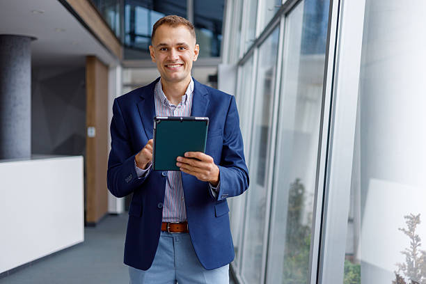istock Confident businessman standing in a contemporary office corridor, looking at camera and working with a tablet device 2239972025