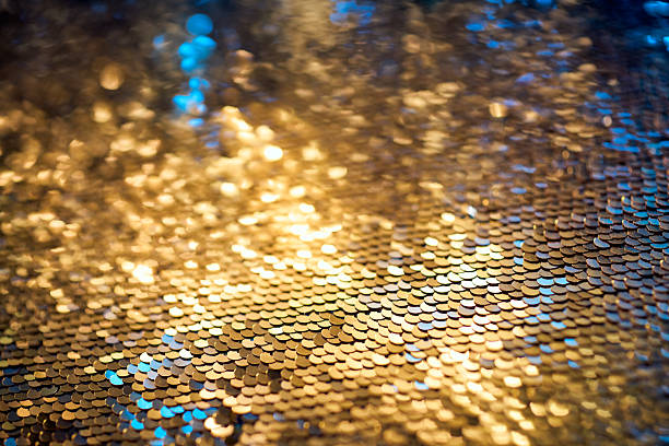 Abstract close-up of a golden sequin surface with glowing reflections and subtle blue highlights. The textured composition evokes festive elegance, luxurious design, and artistic brilliance through vibrant light, soft focus, and metallic shimmer.