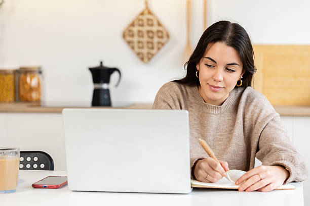 istock Young happy student woman taking notes while using laptop in the kitchen at home 2239975034