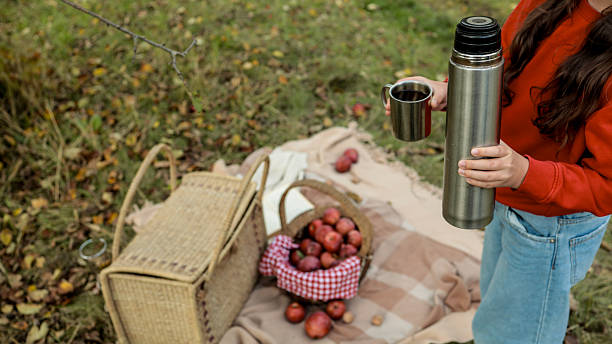 A person in a red sweater and jeans is holding a thermos and a cup of tea during a picnic in the autumn forest. There are apples and a basket.