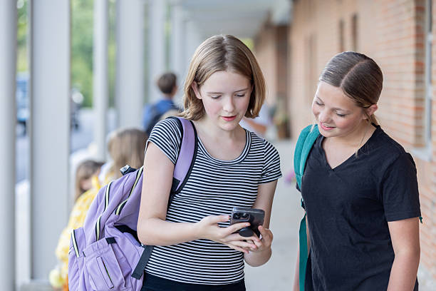 istock Middle school age girls using smart phone look at social media app together while standing outside small town school 2239990064