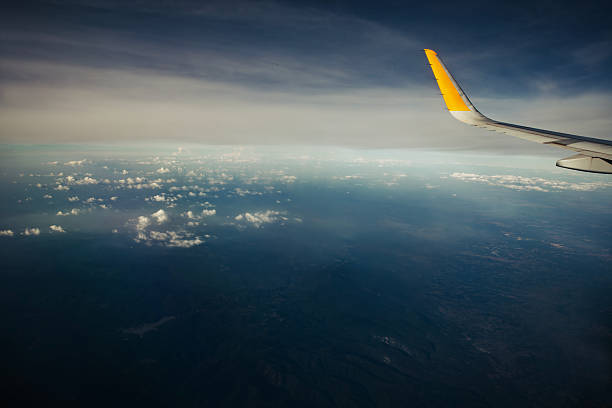 Aerial view from an airplane window, with the aircraft's yellow wing visible against a backdrop of clouds and the sea