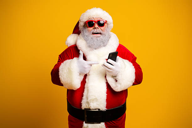 istock Cheerful Santa Claus wearing sunglass using a smartphone against a vibrant yellow backdrop conveying a modern Christmas spirit 2240003687