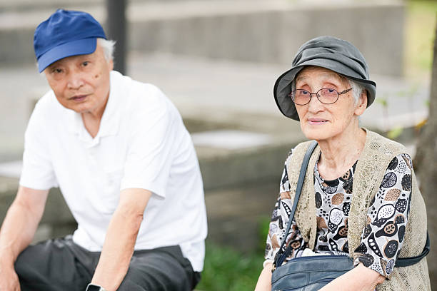 Two elderly people sitting on a bench, one wearing a hat and glasses. The woman is holding a purse