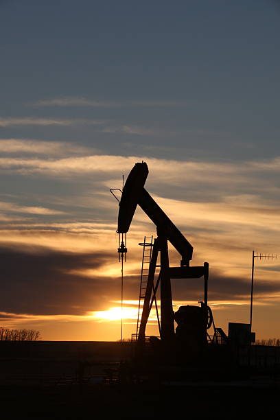 A silhouetted oil pumpjack stands against a vivid Alberta sunset, its mechanical form etched sharply into the sky. Framed by fencing and set on prairie land, the scene captures the intersection of industrial energy production and natural beauty. The warm gradient of the sky contrasts with the stark geometry of the pumpjack, evoking both grit and grandeur.