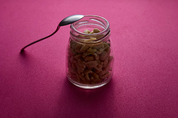 Glass jar of roasted peanuts with a metal spoon balanced on top, photographed on pink background. Healthy natural snack concept with minimalist style and studio lighting.