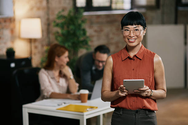 istock Confident professional woman poses with tablet in modern office while colleagues collaborate at table 2240018218
