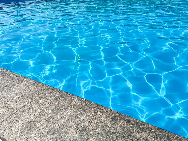 Close up view of vivid blue water in a swimming pool, showcasing the mesmerizing dance of sunlight ripples on the surface. A textured gray concrete coping partially frames the scene, providing a tranquil and inviting atmosphere for relaxation or leisure activities during warm weather. This refreshing liquid background evokes feelings of summer and aquatic refreshment.