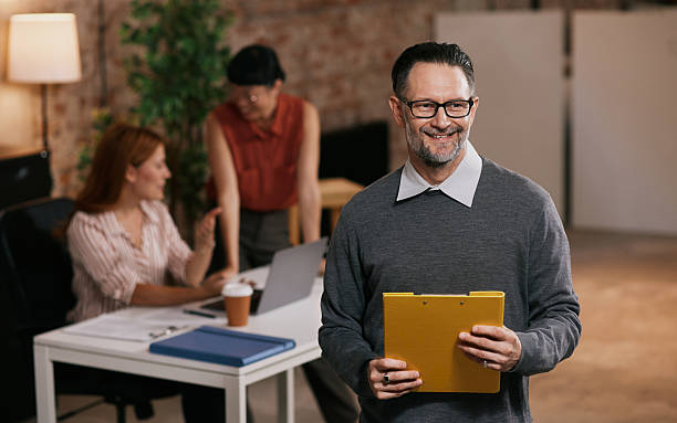istock Professional Man Reading Yellow Folder in Modern Office While Colleagues Work Nearby 2240018500