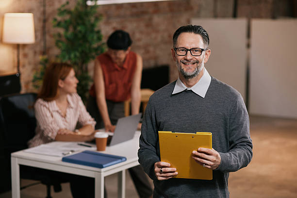 istock Professional Man Reading Yellow Folder in Modern Office While Colleagues Work Nearby 2240018620