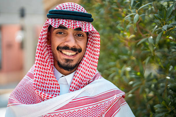 A cheerful young Arabic male in traditional attire smiles warmly outdoors. His keffiyeh and agal contrast beautifully with the greenery around, creating a vibrant, lively scene.