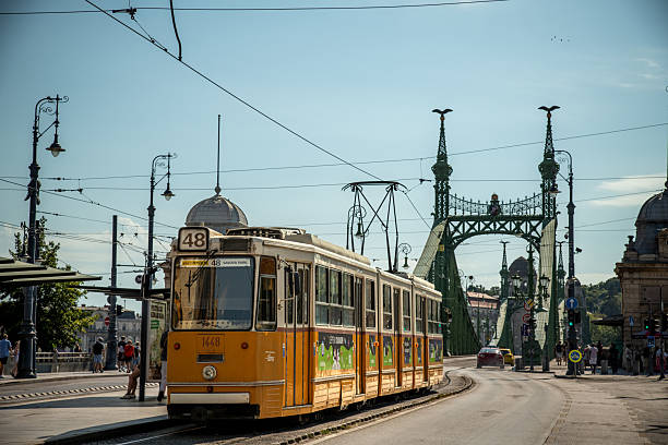 Budapest, Hungary - August 9, 2025: This picture taken in Budapest shows a yellow tram number 48 passing near Liberty Bridge (Szabadság híd). The tram is moving along the street with pedestrians nearby, while the ornate green bridge structure rises in the background under a clear sky.