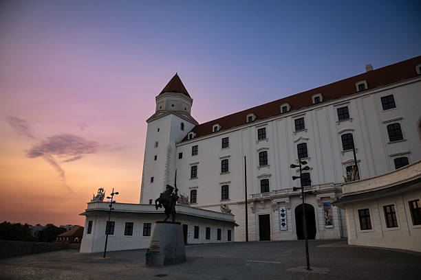 Bratislava, Slovakia - August 6, 2025: This picture taken at Bratislava Castle, Slovakia, shows the castle’s white facade and one of its corner towers under a colorful sunset sky. In the foreground, there is an equestrian statue in silhouette, adding a striking element to the scene.